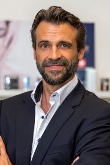 Porträt eines Geschäftsführer Stefan Neider im dunklen Anzug, lächelnd vor einem Regal in Büro- oder Praxisumgebung